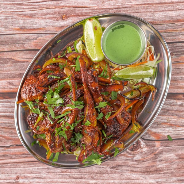 Indian chicken dish with onions, cilantro, lime wedges, and green chutney on a metal platter.