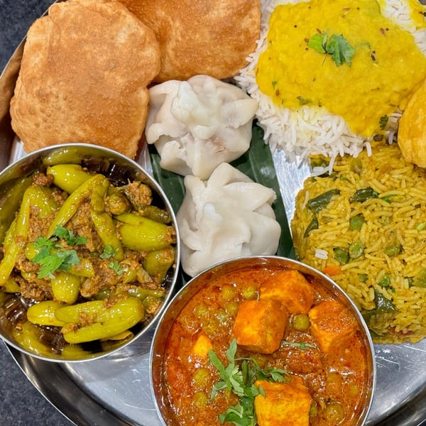 A thali with various Indian dishes: puri, momos, dal, rice, vegetable biryani, paneer curry, and green chili fry.
