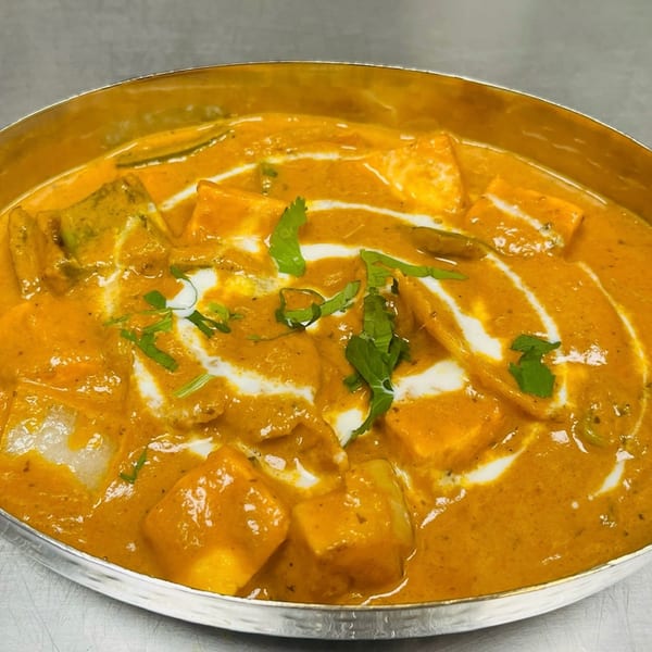 Indian paneer curry with creamy orange sauce, white swirls, and green herb garnish in a silver bowl.